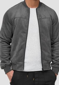 Veste bomber en daim gris avec fermeture éclair à l'avant, poignets côtelés et texture lisse ; portée sur une chemise texturée gris clair.