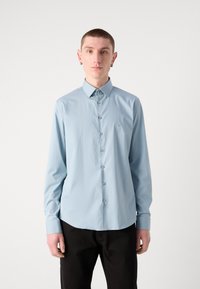Camisa de manga larga de color azul claro, con cuello puntiagudo, textura suave y un patrón sutil. Presenta botones grises y un dobladillo curvado.