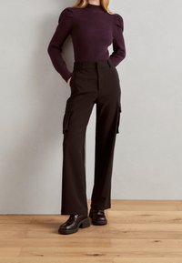 Fialový žebrovaný pletený svetr s nabíranými rameny, doplněný černými kalhotami s vysokým pasem a bočními kapsami, zakončený robustními černými botami.