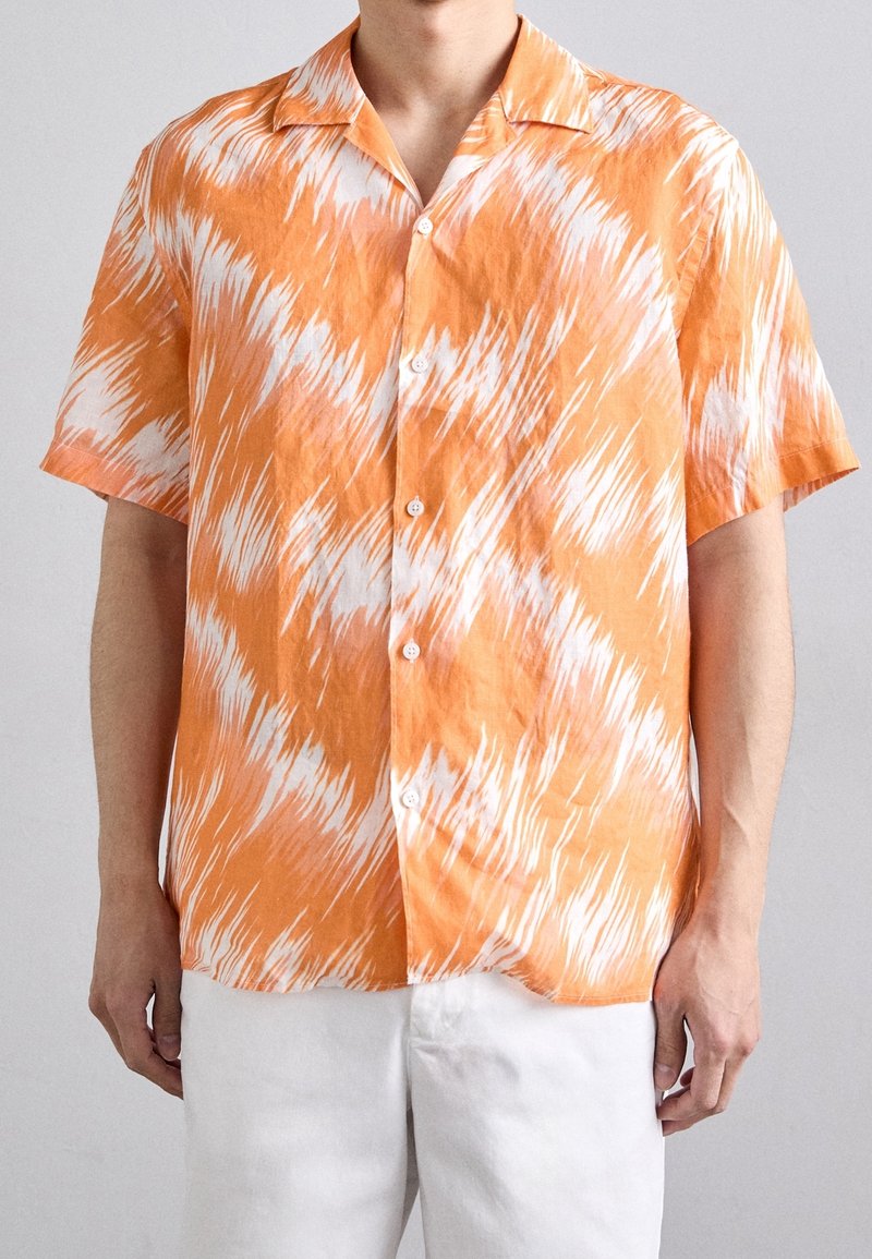 Homme portant une chemise à manches courtes orange et blanche à motifs, boutonnée, avec un pantalon blanc, debout devant un fond uni gris clair.