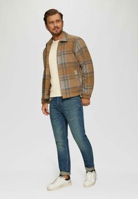 Kariertes Jackett in Braun-, Blau- und Grautönen, mit Reißverschluss und zwei Seitentaschen, kombiniert mit einem cremefarbenen Pullover und blauen Jeans. Weiße Sneakers.