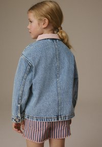 Chaqueta de mezclilla azul claro con un corte relajado, que presenta un collar y acentos de botones, combinada con pantalones cortos a rayas rosas y blancos.