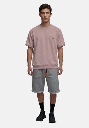 Homme debout, de face, portant un sweat-shirt rose à manches courtes, un short gris avec des cordons orange, des chaussettes noires et des baskets noires.