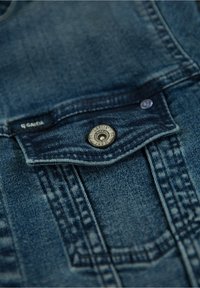 Džínové lacláče s tmavě modrým omakem, které mají přední kapsu, kovové zapínání na knoflík a prošívané detaily podél švů.