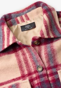 Chemise en laine à carreaux avec des couleurs rouge, bleu et crème. Présente un col pointu, une fermeture à boutons et une étiquette à l'intérieur. Texture douce.