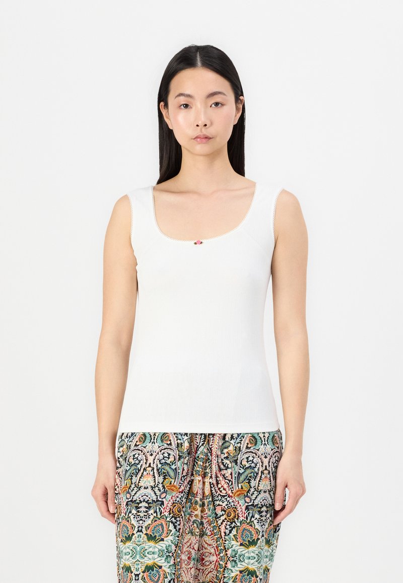 Top de tirantes blanco con un escote cuadrado, que presenta un pequeño acento floral en el centro. Llevado con pantalones estampados y coloridos.