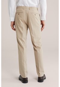 Mann trägt beige Anzughose mit Gesäßtaschen und braune Anzugschuhe, steht vor einem schlichten hellen Hintergrund.