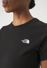 Primo piano di una persona che indossa una maglietta nera a girocollo con il logo bianco di The North Face sul lato sinistro del petto.