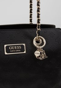 Sac à main synthétique noir avec des motifs en relief, une bandoulière en chaîne dorée et des pendentifs décoratifs de la marque avec une étiquette logo.