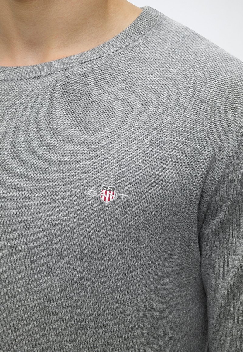Close-up of a person wearing a grey knit sweater with a small shield emblem and white text on the chest.