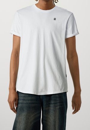 Personne portant un t-shirt blanc uni à manches courtes avec un petit logo noir sur le haut gauche de la poitrine, assorti à un jean en denim bleu foncé.