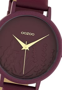 Montre bourgogne avec cadran texturé présentant des motifs de plantes en relief ; aiguilles dorées et logo de la marque. Bracelet en matériau doux et boîtier rond.
