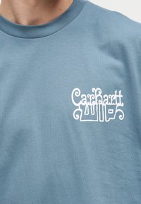 Nahaufnahme einer Person, die ein blaues Sweatshirt mit einem weißen stilisierten „Carhartt WIP“-Logo auf der linken Brust trägt.