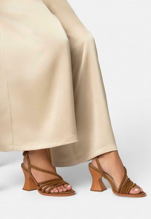 Piedi che indossano sandali marroni con tacco e più cinturini, abbinati a pantaloni beige a gamba larga, su sfondo bianco semplice.