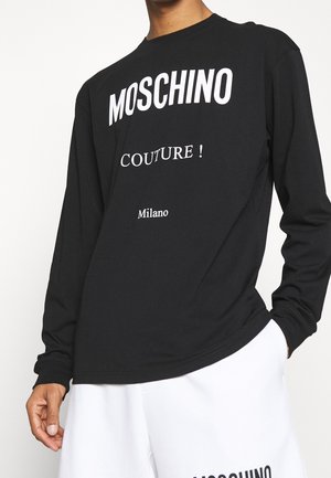 Schwarzes Langarmshirt mit weißem "MOSCHINO COUTURE! Milano"-Schriftzug, getragen zu weißen Shorts, Hand ruht an der Hüfte.