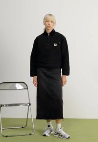 Schwarze gepolsterte Jacke, schwarze Midirock, weiße Turnschuhe mit gelben Akzenten. Durchsichtiger Stuhl auf einem grünen Boden mit schlichtem weißen Hintergrund.