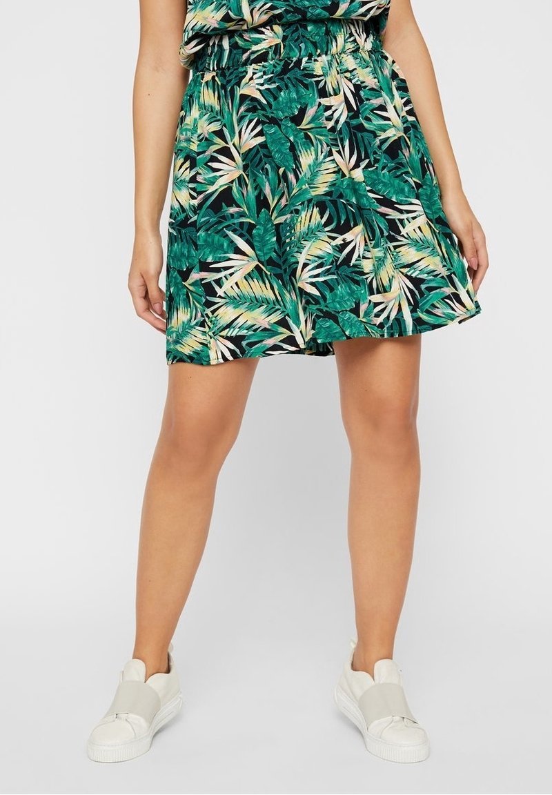 Femme portant une jupe imprimée de feuilles tropicales vertes avec une taille élastique et des baskets blanches à enfiler sur un fond uni.