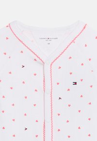 Λευκό βρεφικό φορμάκι με ροζ σχέδια καρδιάς, ροζ διακοσμητικό περίγραμμα με κυματιστό τελείωμα κατά μήκος των κουμπιών κ αι λογότυπο Tommy Hilfiger στο στήθος.