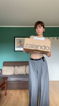 Kartonová krabice Zalando, světle hnědá s šedým logem. Modelka má na sobě bílý krátký top a šedé široké kalhoty, drží krabici.