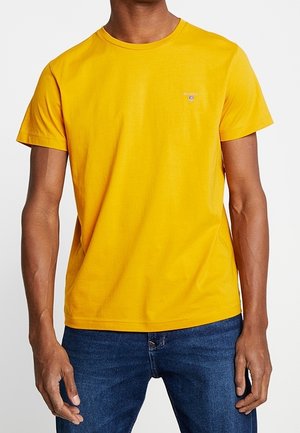 T-shirt en coton jaune avec col rond et manches courtes, arborant un petit logo sur la poitrine. Associé à un jean en denim bleu.