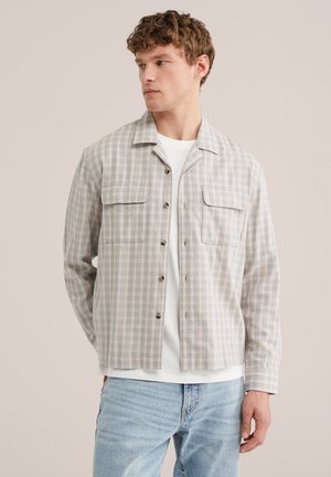 Młody mężczyzna w beżowej koszuli w kratę zapinanej na guziki, na białej koszulce i jasnoniebieskich dżinsach, stojący na jednolitym tle.