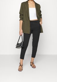 Blazer vert olive sur un crop top blanc, associé à un pantalon noir taillé et des sandales noires à lanières. Petit sac à main noir avec une texture matelassée.