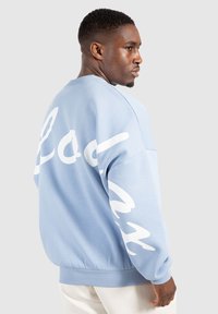 Lichtblauwe sweatshirt met lange mouwen, voorzien van grote witte cursieve letters op de achterkant. Ribbelmanchetten en -zoom. Glad textuur.