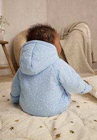 Jasnoniebieska hoodie w gwiazdki z kieszenią kangurzą i miękką teksturą, noszona przez niemowlę siedzące na pikowanej kocyku.