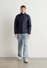 Navy puffer jacket with horizontal quilted pattern, full zip, and collar, paired with light blue denim jeans and white sneakers.
