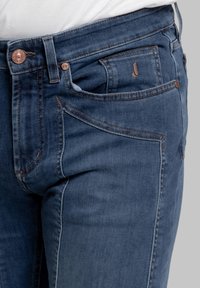 Vaqueros de mezclilla en un lavado azul oscuro con un ajuste slim. Las características incluyen costuras en contraste, dos bolsillos frontales y un bolsillo trasero con un detalle de logotipo.