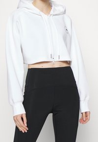 Sudadera blanca corta con cremallera y capucha, de mangas largas y con un logo. Combinada con leggins negros ajustados, que presentan una textura suave.
