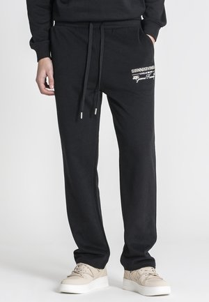 Hombre que lleva pantalones deportivos negros con texto de logotipo blanco en el muslo, zapatillas beige y sudadera negra con una mano en el bolsillo.