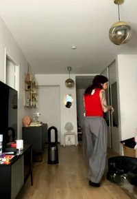Άτομο με κόκκινο αμάνικο τοπ και γκρι παντελόνια περπατά σε ένα μοντέρνο σαλόνι με ξύλινο πάτωμα, μαύρο σταντ τηλεόρασης, καθαριστή αέρα και κρεμαστά φωτιστικά.