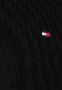 Tissu tricoté noir avec un petit logo brodé comportant des rectangles rouges et blancs sur le côté gauche. Texture lisse.