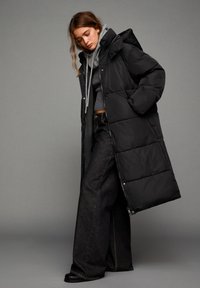 Cappotto lungo nero imbottito con cappuccio, tessuto trapuntato e zip laterale, abbinato a una felpa grigia e jeans larghi scuri.