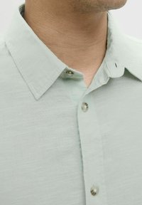 Camisa de color verde claro con cuello hecho de tejido texturizado, que presenta botones cosidos y un cuello abotonado. Primer plano del escote y la parte superior del pecho.