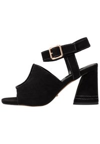 Svart mocka öppen tå sandal med blockklack, ankelrem med guldfärgad spänne och vita sömnadsdetaljer.