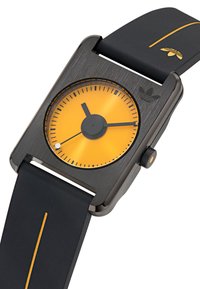 Montre carrée avec un boîtier métallique noir et un cadran jaune. Présente un bracelet en caoutchouc noir avec une bande jaune et un logo, ainsi que des aiguilles rondes pour les heures et les minutes.