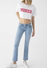 Bílý crop top s nápisem „INDEED“ tučnýma růžovýma písmeny, doplněný světle modrými džínami a bílými teniskami.