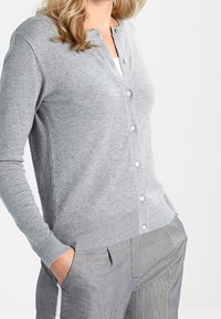 Kvinna klädd i en ljusgrå knappad kofta med subtila strukturerade prickar och grå rutiga byxor, med händerna avslappnat i fickorna.