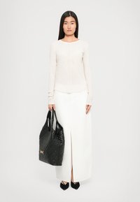 Woman wearing a long white skirt with front slit, cream long-sleeve top, black pointed shoes, holding a large black quilted handbag.