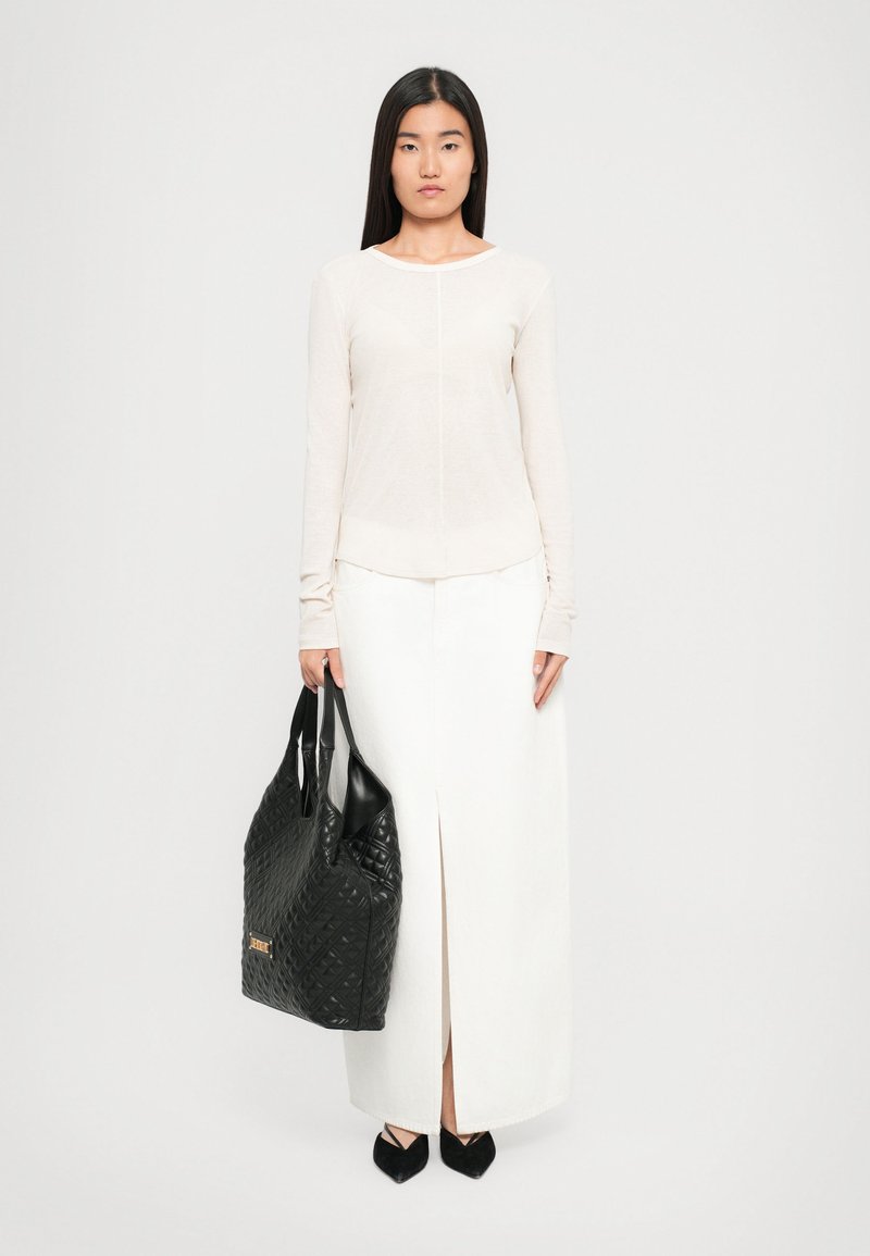 Woman wearing a long white skirt with front slit, cream long-sleeve top, black pointed shoes, holding a large black quilted handbag.