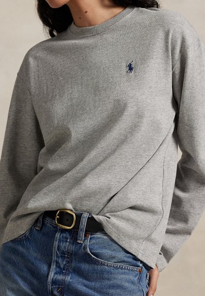 Donna che indossa una maglietta grigia a manica lunga con un logo blu ricamato, infilata in jeans blu con una cintura nera e fibbia dorata.