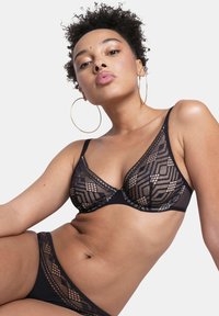 Ensemble de lingerie en dentelle noire avec des motifs géométriques, un design transparent et un tissu uni contrastant. Bretelles réglables et détails décoratifs présents.