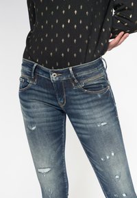 Jeans de mezclilla en un lavado azul oscuro con parches desvaídos y detalles desgastados. Cuenta con un diseño clásico de cinco bolsillos y cierre de botón.