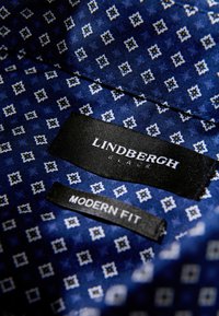 Blauw patroonstof met kleine witte bloemen, voorzien van zwarte labels met de tekst "LINDBERGH BLACK" en "MODERN FIT." Gladde textuur.
