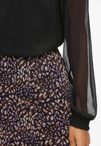 Blusa nera con maniche lunghe trasparenti abbinata a una gonna a fantasia marrone e viola realizzata in un tessuto leggero e fluido.