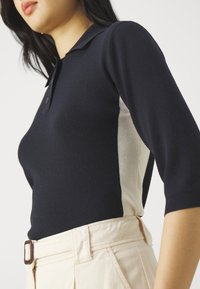 Marine gebreide polo met korte mouwen, voorzien van lichtcrème zijpanelen. Gekraagde stijl met knopen en een soepele textuur. Draagt met beige broeken.