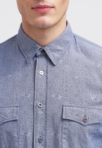 Light blue patterned shirt with a button-down collar, two chest pockets, and light-colored buttons. The fabric has a subtle texture.