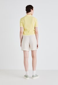 Gul, rillet kortermet polo med hvite detaljer, kombinert med beige shorts. Modellen har på seg grønne joggesko og hvite sokker. Enkel bakgrunn.